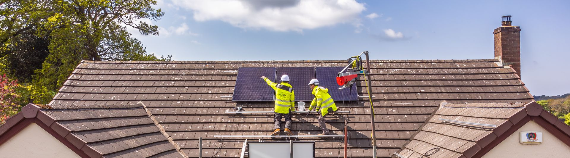 Two workers in high-visibility yellow jackets install solar panels on a brown tiled roof on a sunny day.
