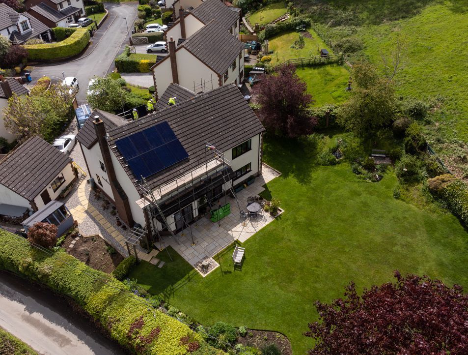 An aerial view of a two-story house with solar panels on the roof, featuring a patio and a large green lawn.