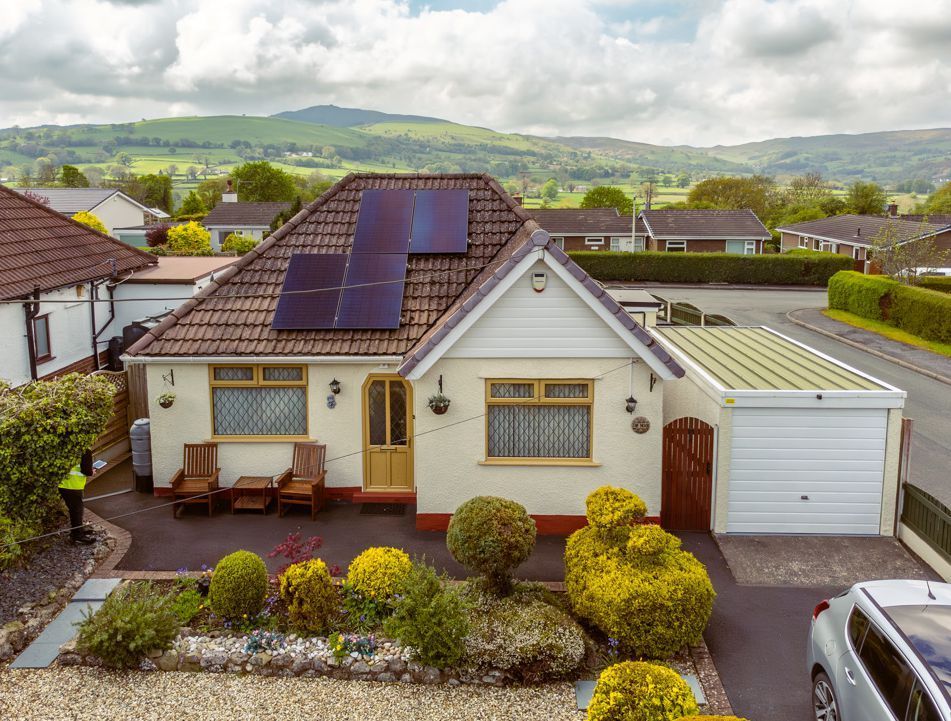 A cream-colored bungalow with solar panels on the roof, a white garage, and a landscaped front garden with mountain views.