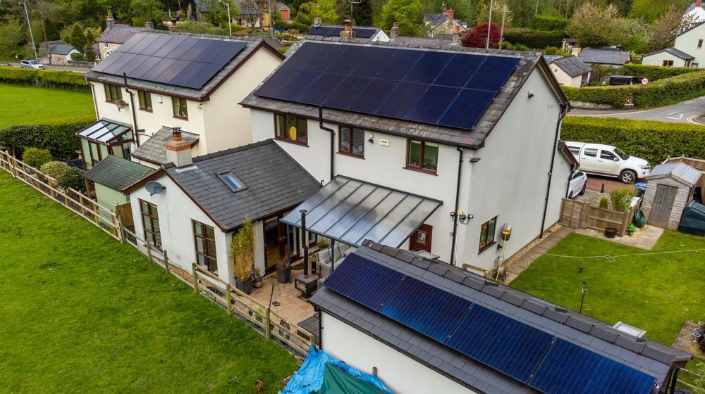 An aerial view of two connected houses in a rural setting, both featuring prominent solar panel arrays on their roofs.