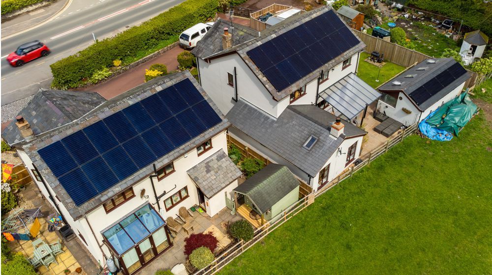 Aerial view of two white houses with dark solar panels on their roofs, located next to a road and a green grassy field.