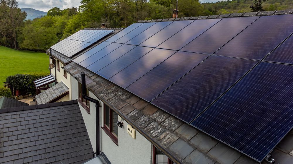 Black solar panels installed on a residential roof in a scenic, grassy rural area.