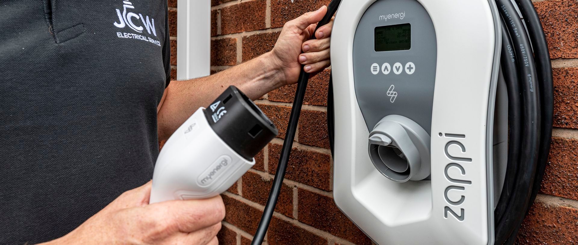 A person holds a Type 2 electric vehicle charging cable near a wall-mounted white zappi EV charger on a brick wall.