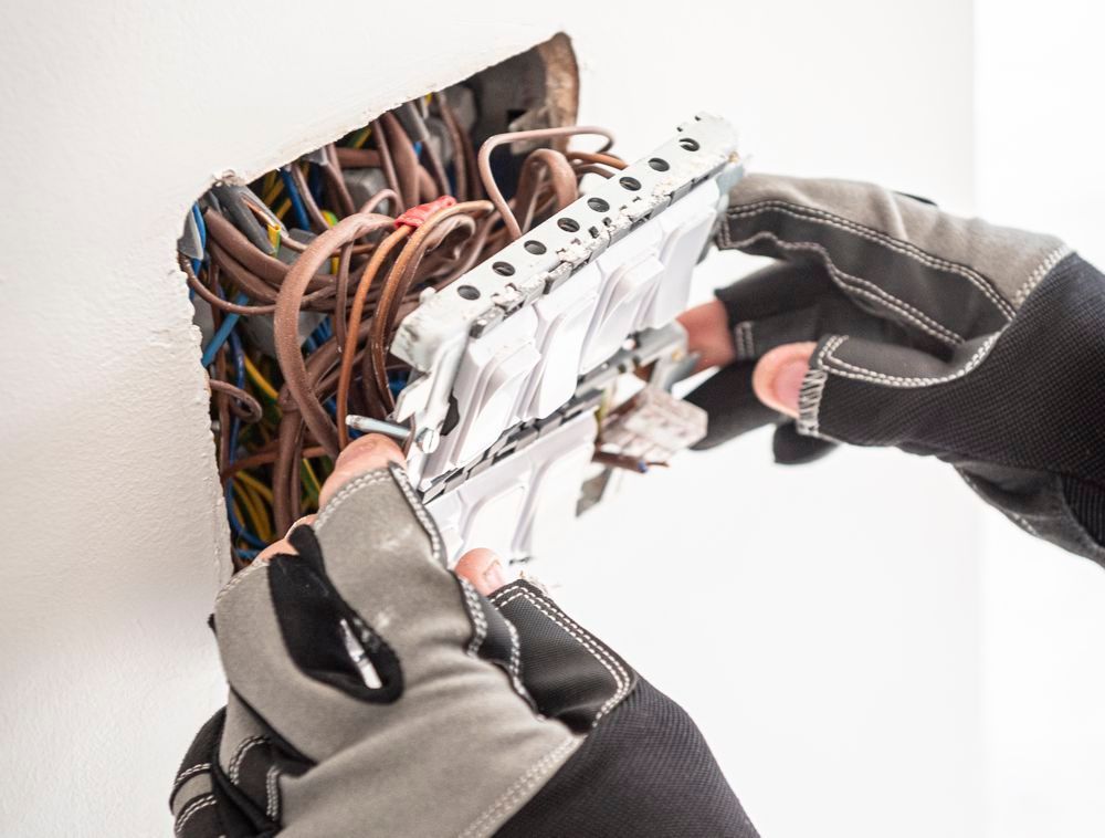 A person wearing gloves pulls a light switch assembly from a wall junction box filled with electrical wiring.
