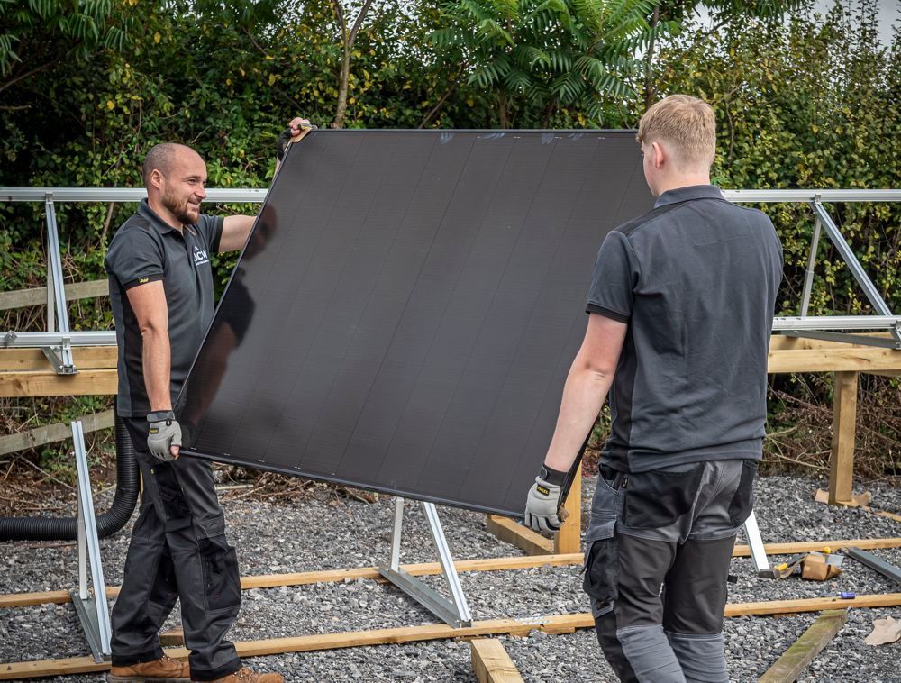 Two people wearing work clothes carry a large, square black solar panel outdoors on a construction site.