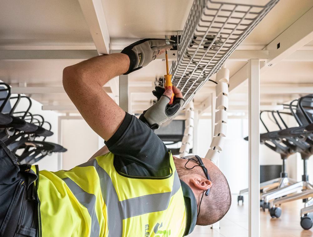 A person wearing a high-visibility vest and gloves uses a screwdriver to work on electrical wiring in an office space.