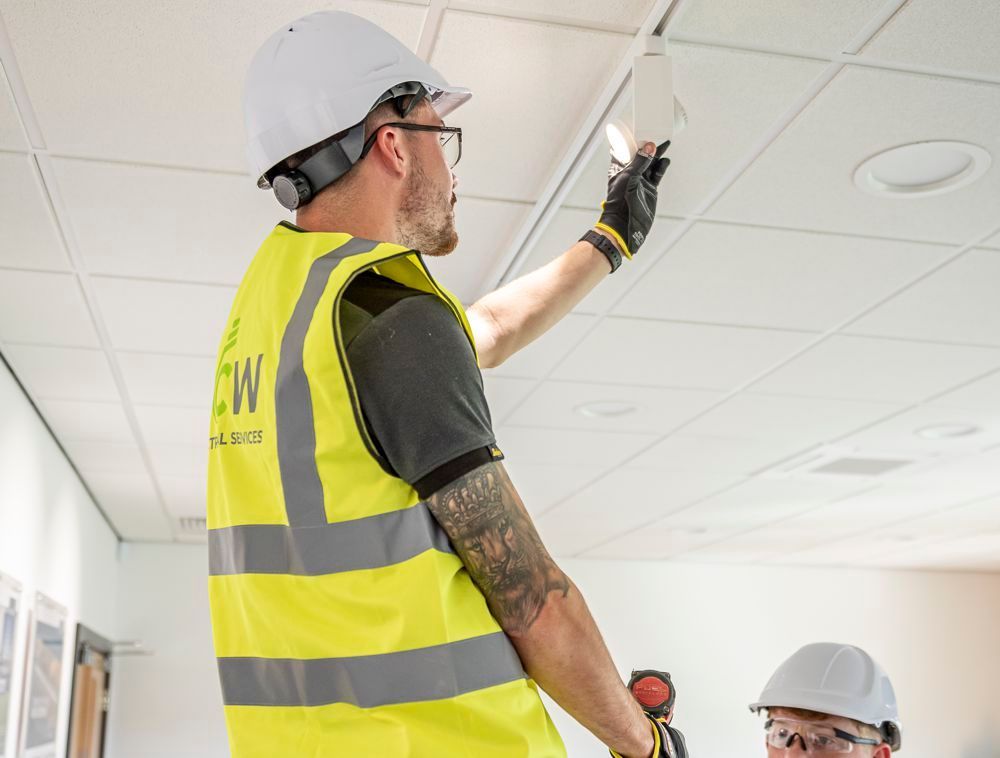 Two workers in hard hats and high-visibility vests work on installing a light fixture in a drop ceiling.