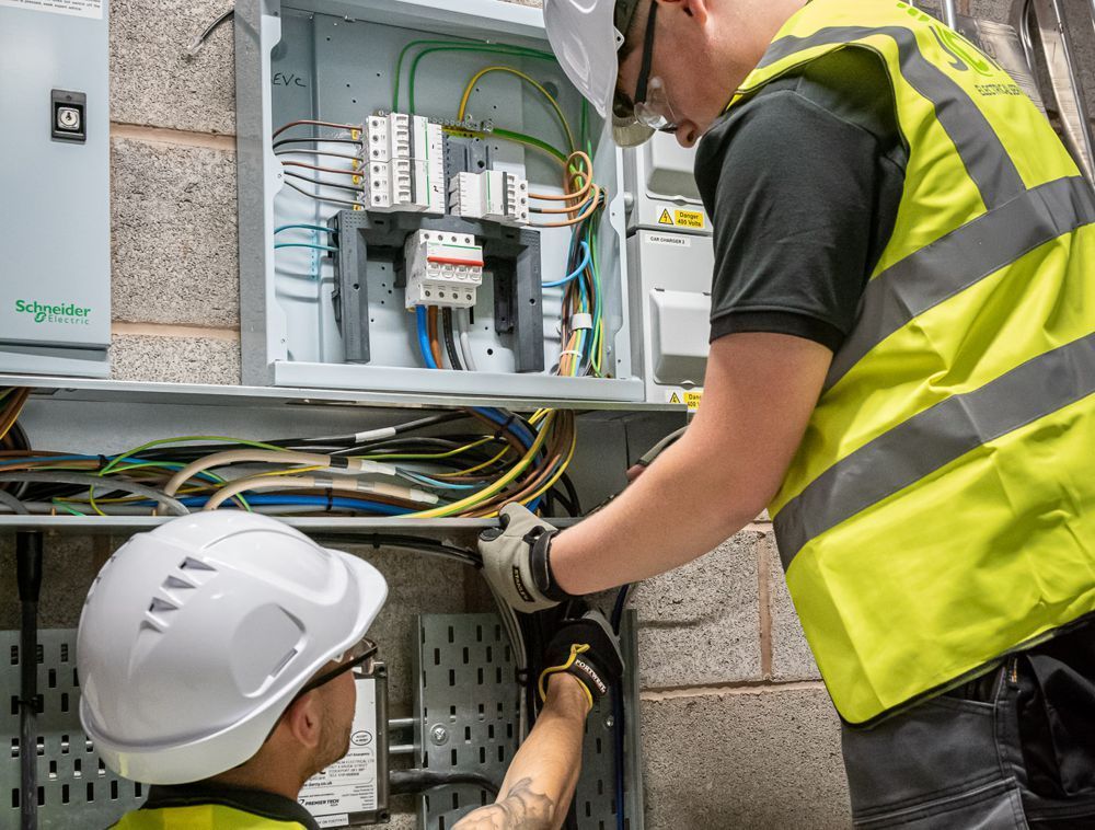 Two workers wearing hard hats and high-visibility vests perform electrical work on a circuit board mounted on a brick wall.