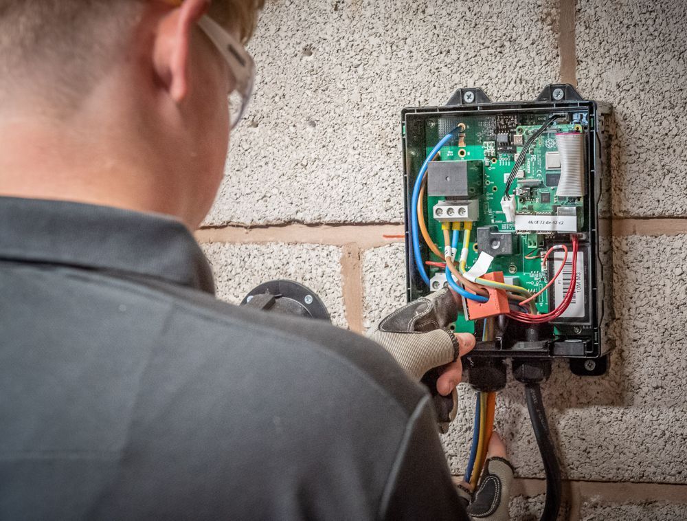 A person wearing protective eyewear is installing or repairing electrical wiring inside a wall-mounted control unit.
