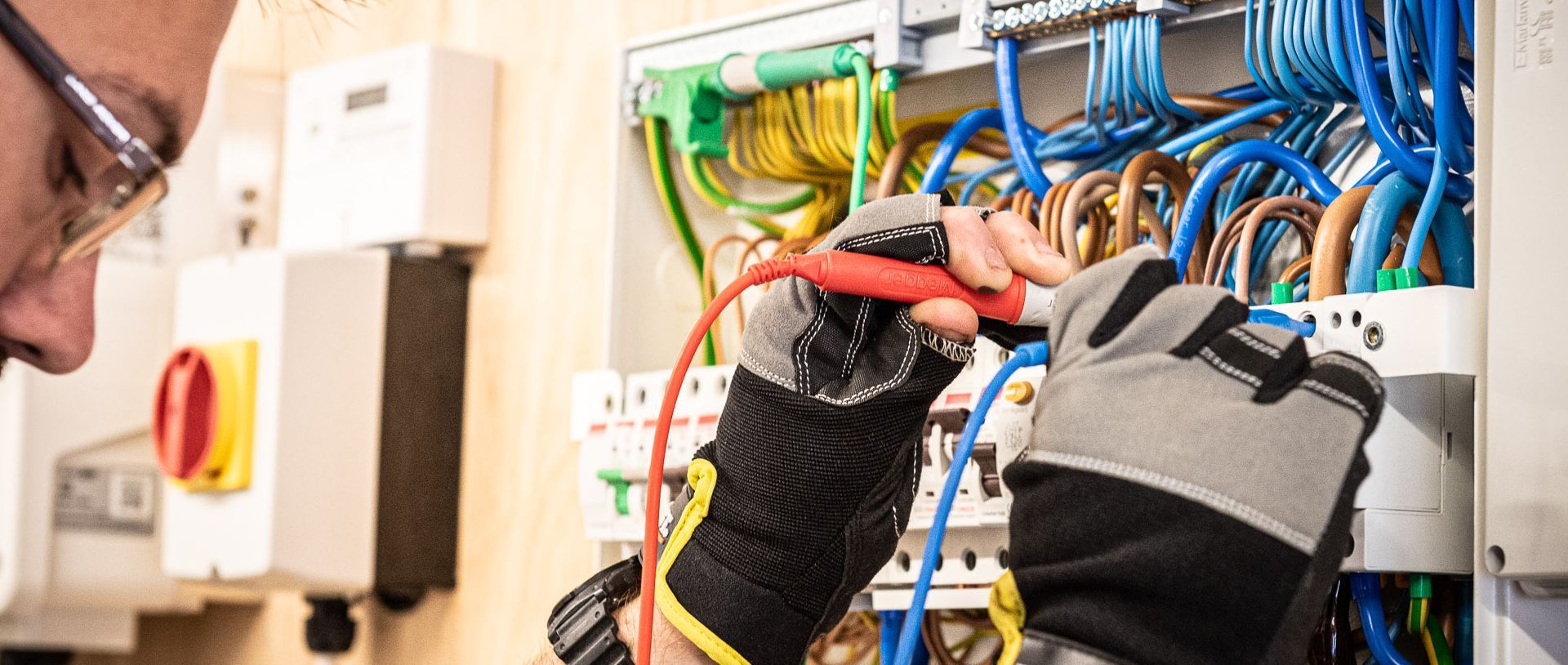 An electrician wearing safety glasses and gloves uses a multimeter probe to test a wire inside a circuit breaker panel.