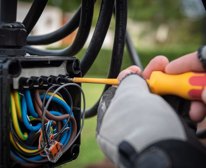 A gloved hand uses a yellow-handled screwdriver on a black electrical junction box filled with multicolored wires.