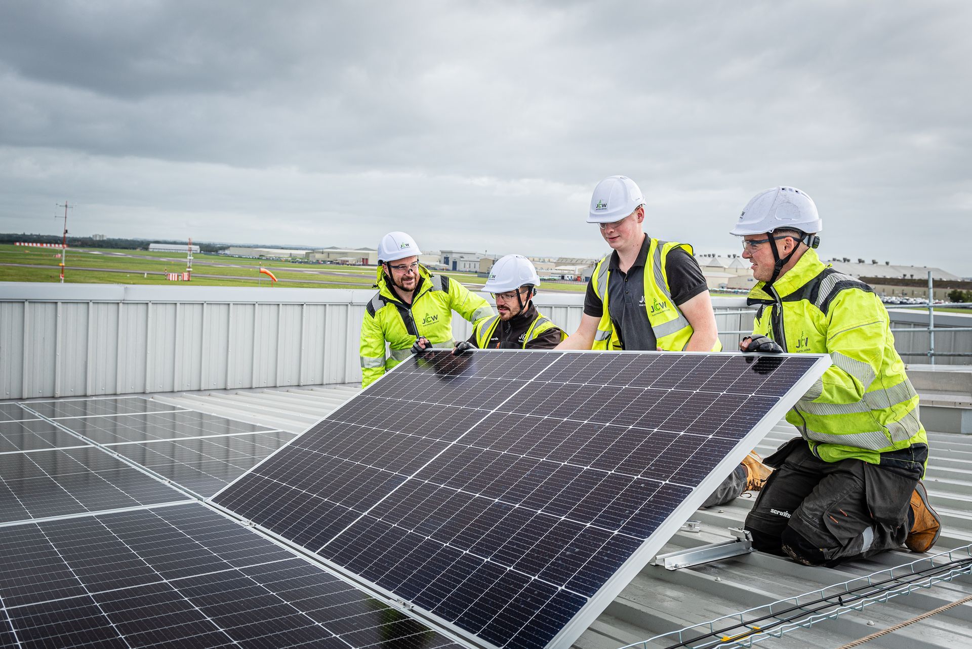 Four people in hard hats and high-visibility vests work together to install a large solar panel on a rooftop.
