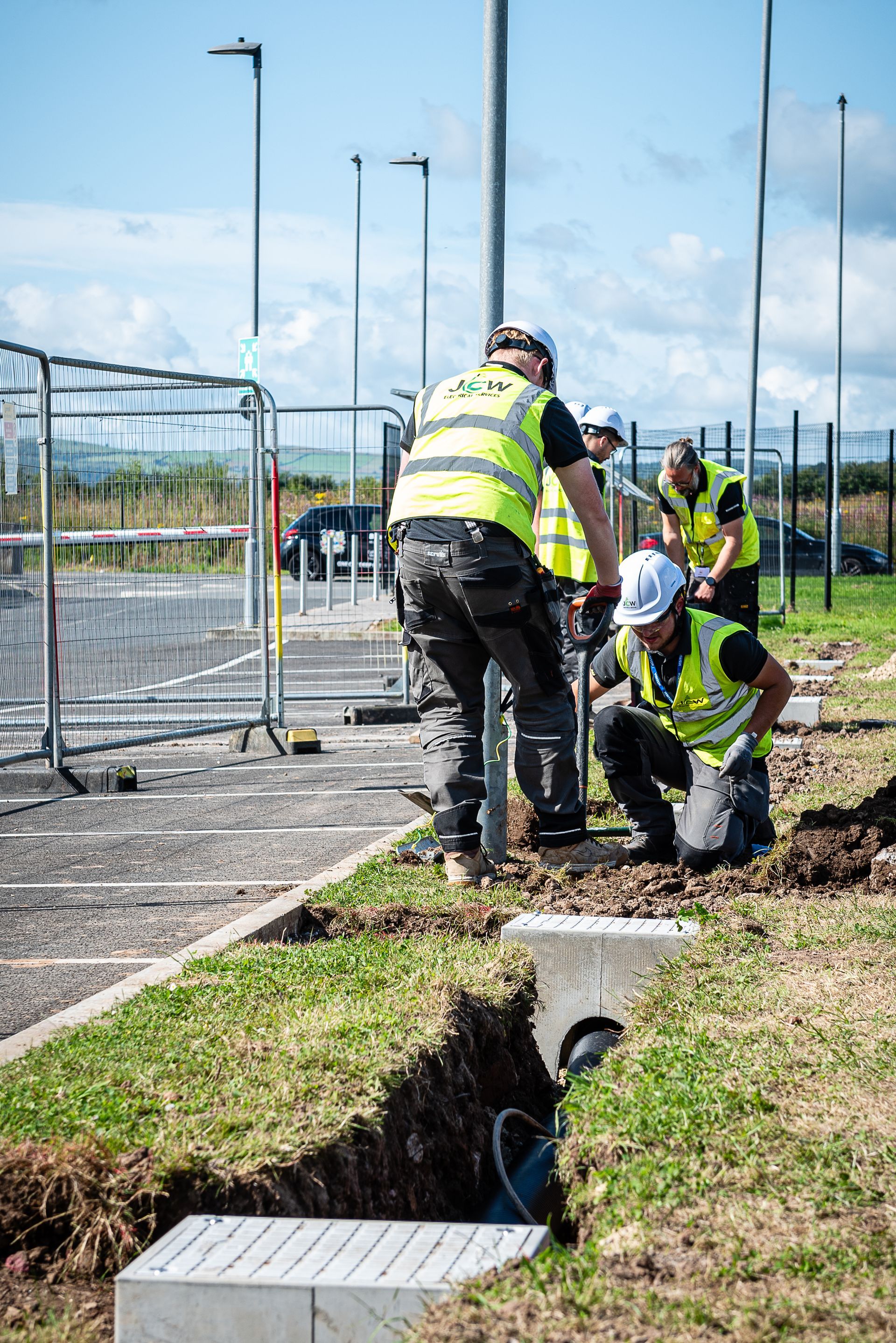 Construction workers in high-visibility vests work on a post and trench in an outdoor lot on a sunny day.