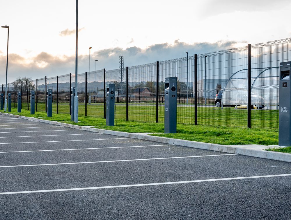 A line of electric vehicle charging stations installed along a parking lot next to a high fence at sunset.