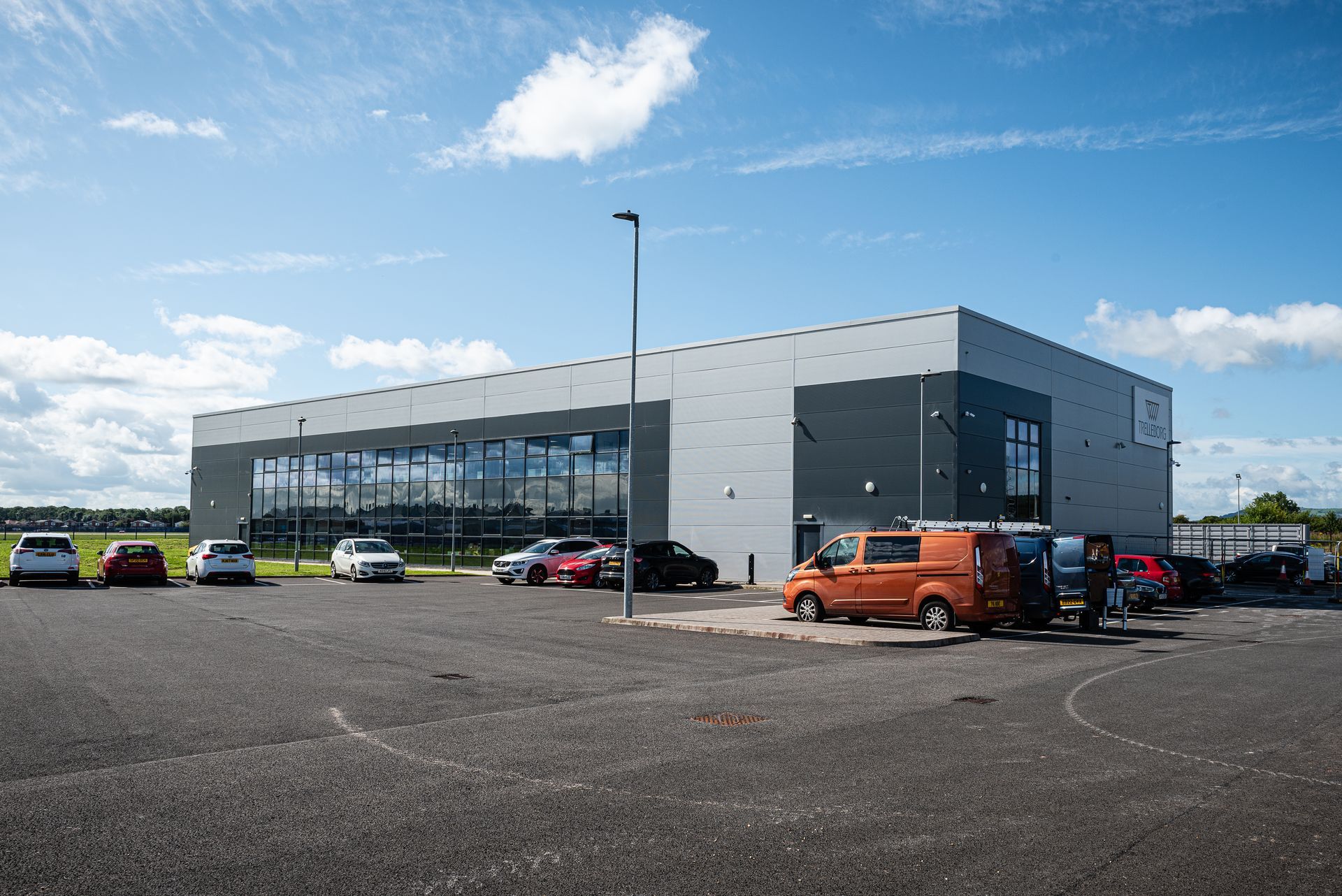 A modern, rectangular industrial building with large windows and gray paneling, set in a parking lot under a blue sky.