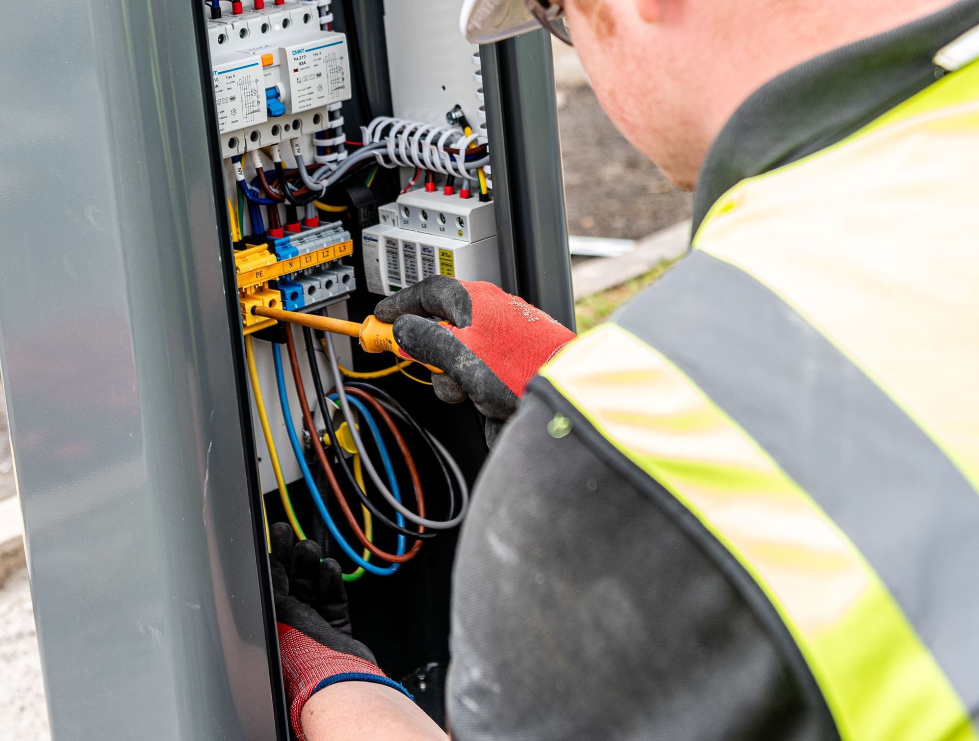 A technician in high-visibility gear uses a screwdriver on electrical components inside an open equipment cabinet.