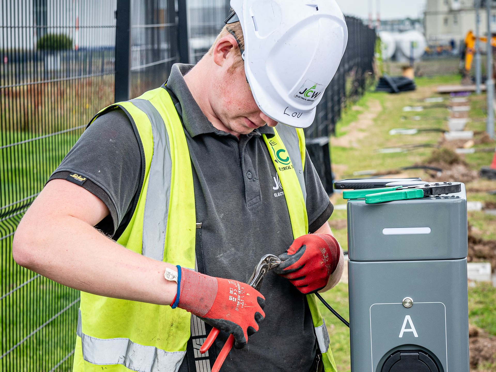 Workers in high-visibility vests perform electrical work on a grassy strip beside a paved parking area and metal fence.