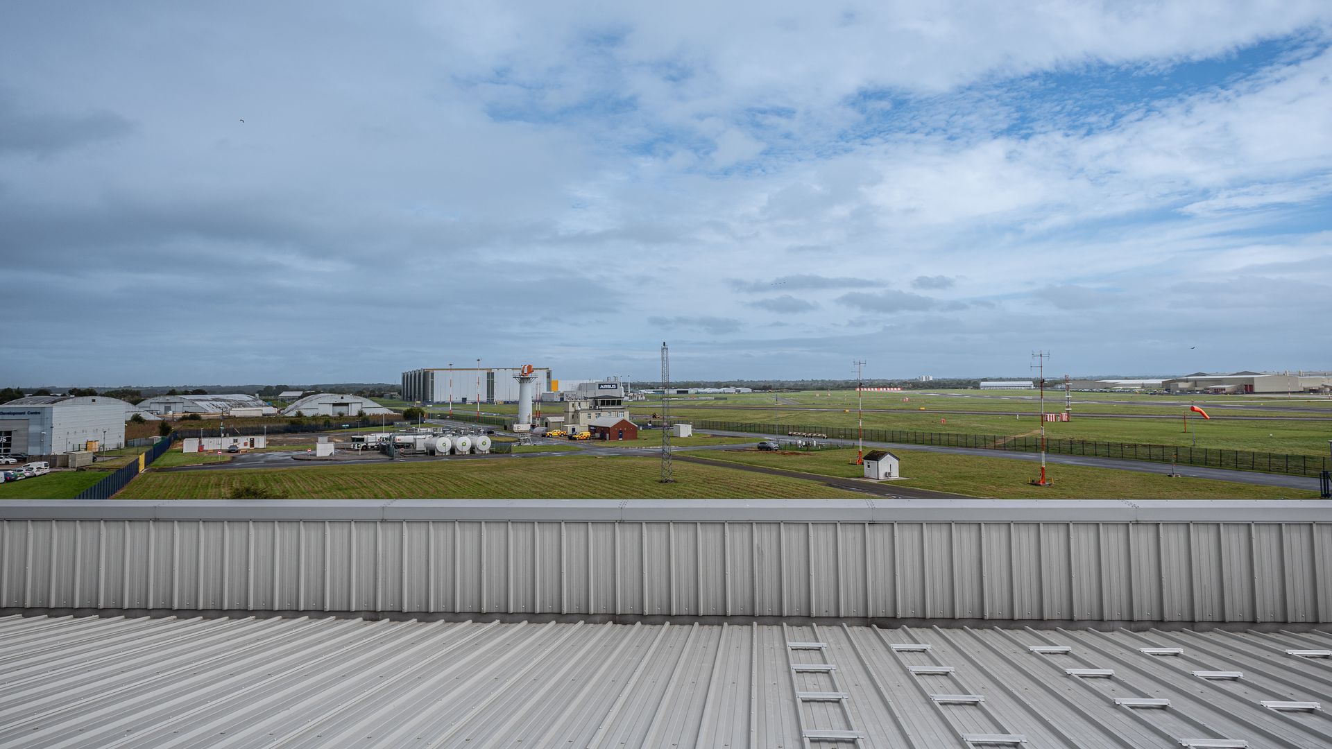 A view from a corrugated metal roof overlooking a grassy industrial area with warehouses under a cloudy blue sky.