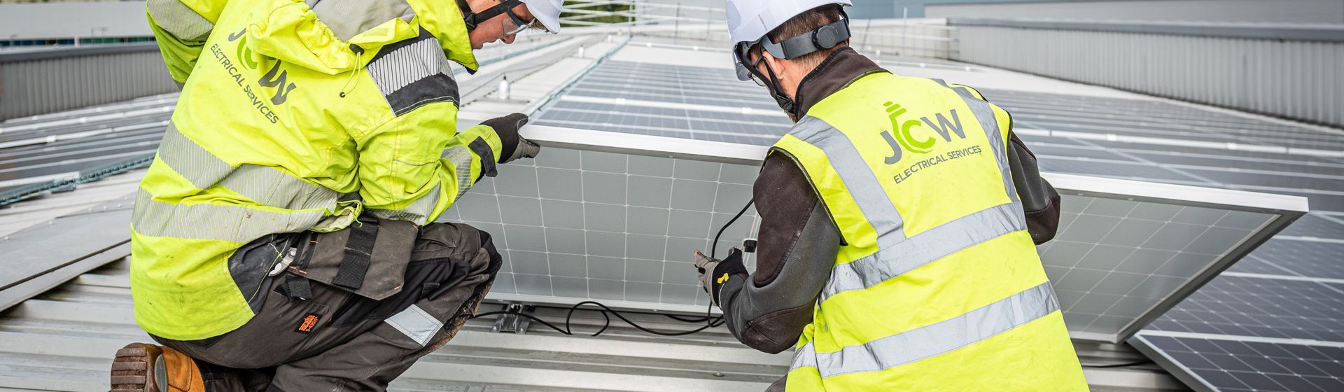 Two workers in high-visibility jackets and hard hats kneeling on a rooftop to install solar panels.
