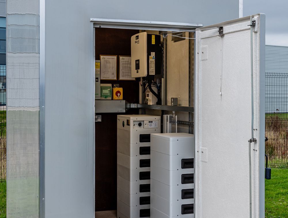 An open outdoor enclosure reveals an industrial solar power inverter and modular battery storage system.