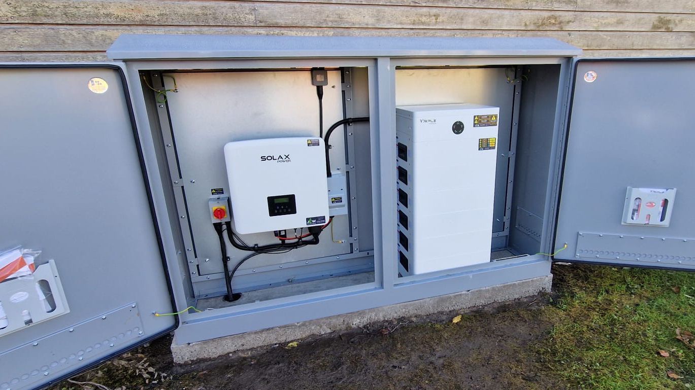 An outdoor electrical cabinet with two open doors, housing a white solar inverter and a white battery storage unit.