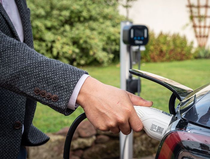 A person in a gray blazer plugs a charging cable into an electric vehicle at an outdoor charging station.