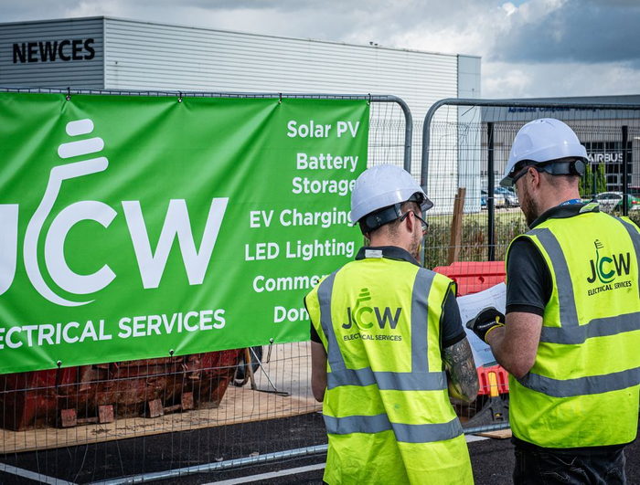 Two professionals in high-visibility vests and hard hats stand before a JCW Electrical Services banner at a construction site.