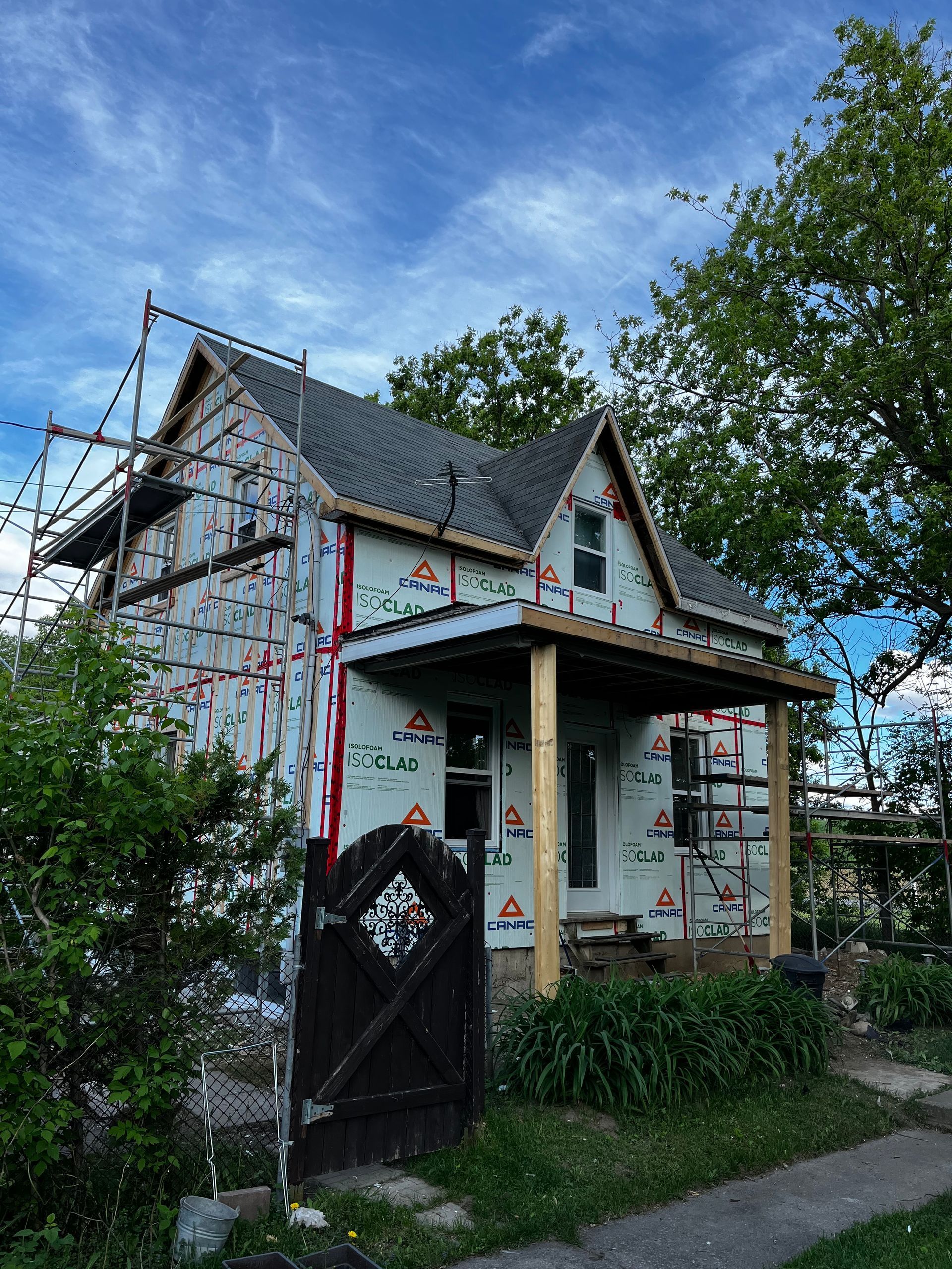 Maison en construction, recouverte d'une bâche Tyvek, échafaudages sur le côté, petit porche, ciel nuageux.