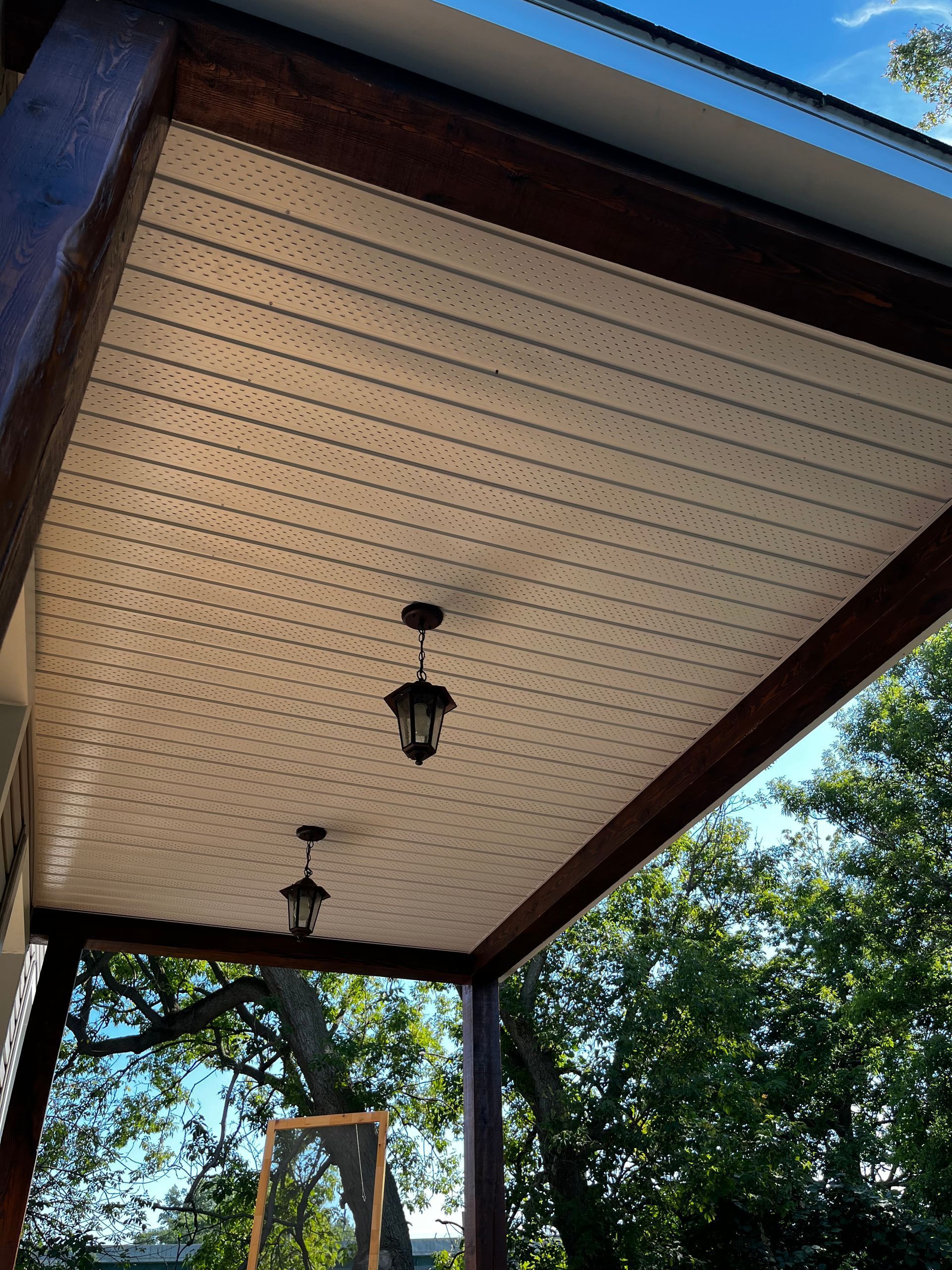 Plafond de la véranda couverte avec moulures en bois foncé, panneaux blanc cassé, deux lampes suspendues et verdure.