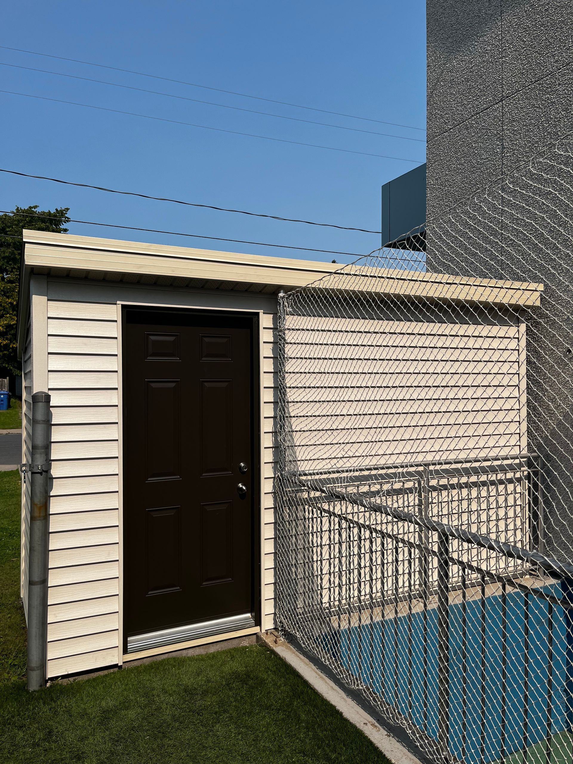 Abri de jardin blanc avec porte marron, rambarde en métal et base bleue, adossé à un mur texturé sous un ciel bleu.