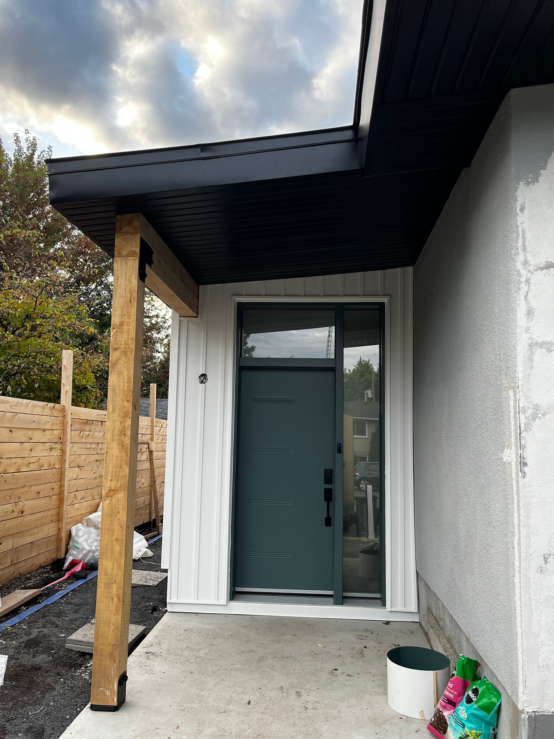 Porte verte avec imposte sous un porche aux boiseries sombres, poteau en bois à gauche, mur en stuc à droite.