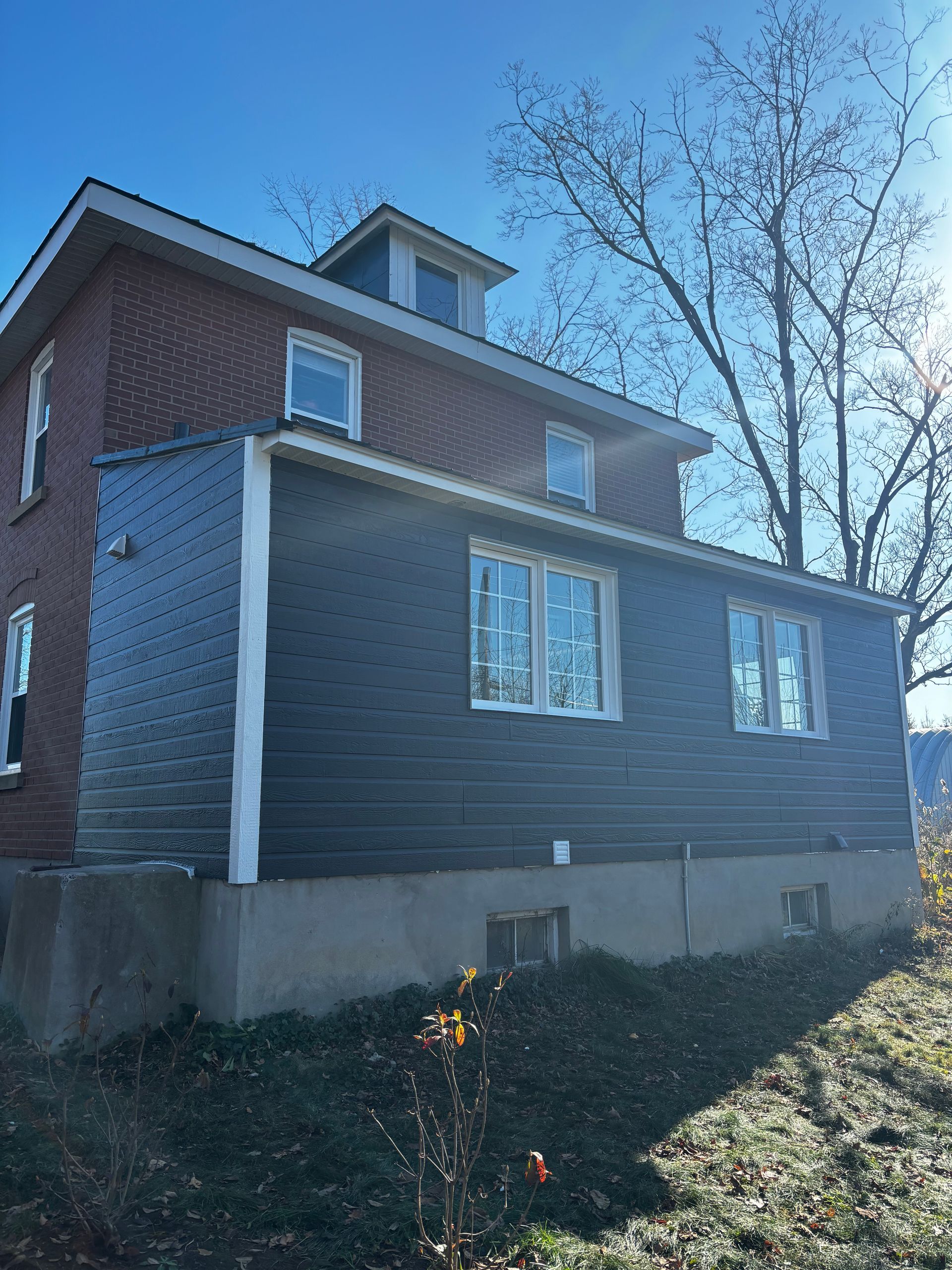 Maison à deux étages, étage supérieur en briques avec bardage foncé, entourée d'arbres sous un ciel bleu.