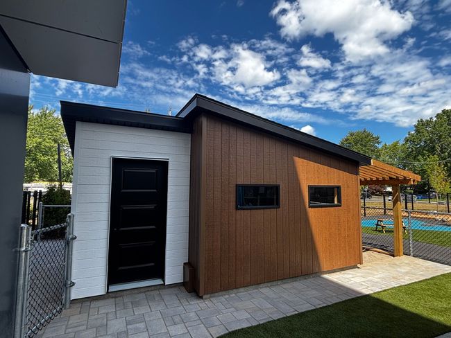 Abri de jardin moderne avec bardage blanc et marron, porte noire et petites fenêtres, sur une terrasse en briques sous un ciel bleu.