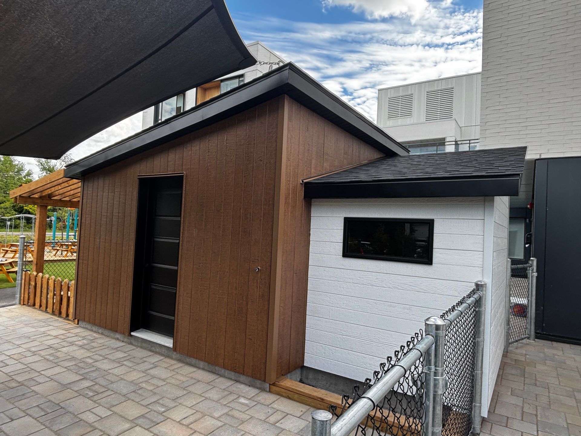 Abri de jardin marron et blanc avec une porte et une fenêtre noires, à côté d'un bâtiment, d'une zone pavée et d'une clôture.