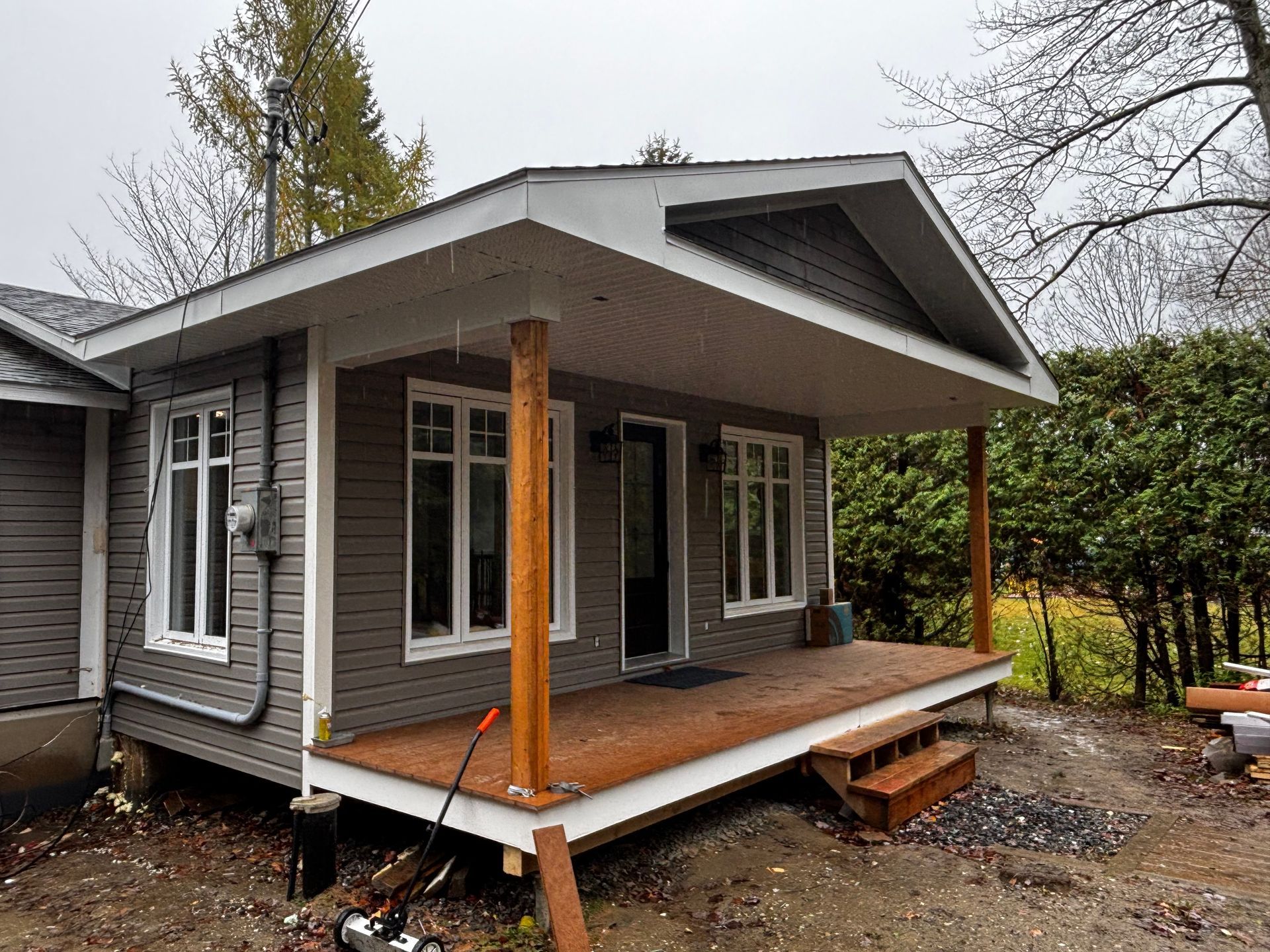 Maison grise avec porche couvert, terrasse en bois, deux marches et poutres de soutien en bois.