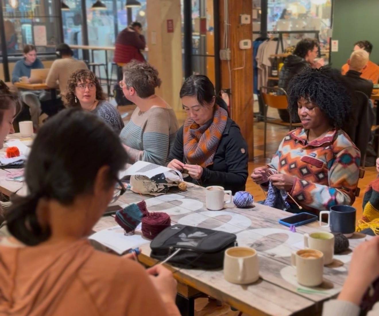 People seated around a table knitting, some sipping from mugs. Setting: a workshop with exposed beams and string lights.