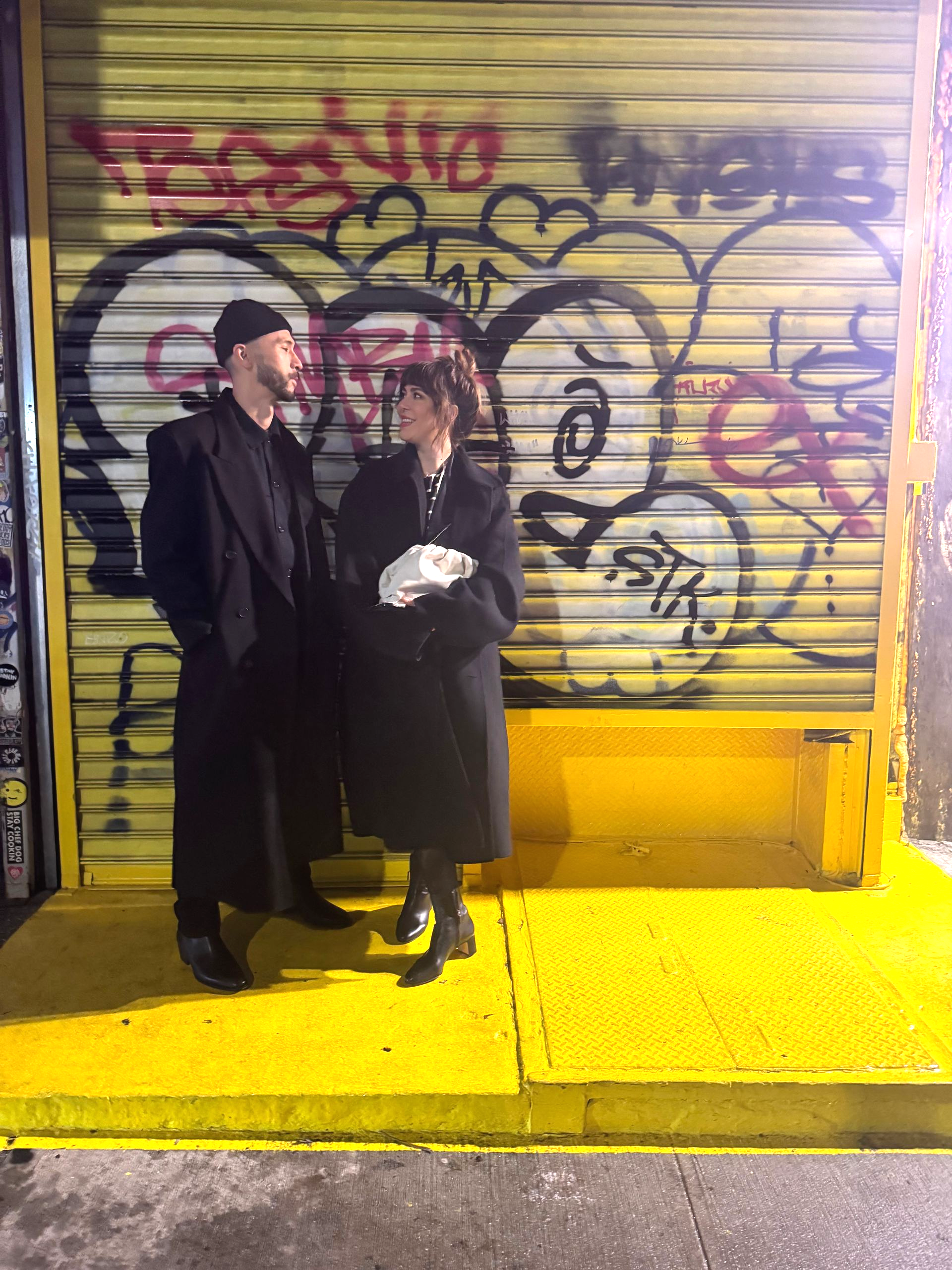 Two figures in black coats stand on a yellow platform in front of a graffiti-covered metal door.