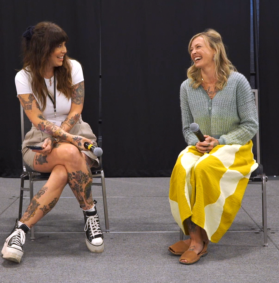 Two women sit on chairs, laughing during a panel discussion. One has tattoos, the other wears a sweater and skirt.
