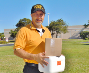 Man in orange shirt holding a box and white container outdoors on a grassy lawn, wearing a cap