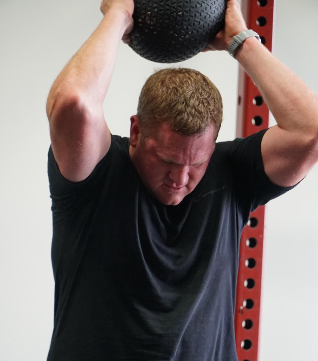 Man in black shirt holding medicine ball overhead, exercising in a gym.