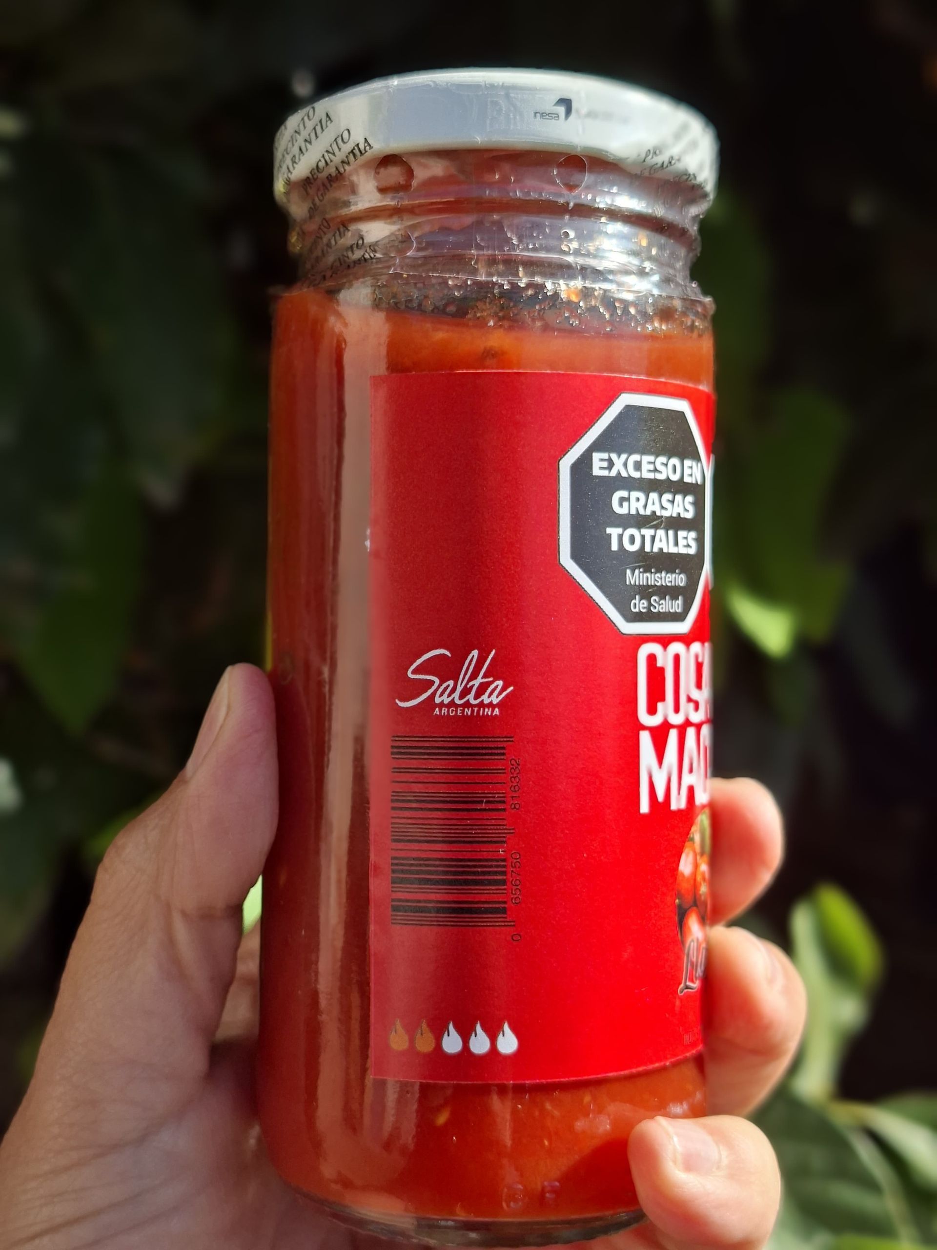 Mano sosteniendo un frasco de salsa roja con tapa blanca y etiqueta roja sobre un fondo oscuro borroso.