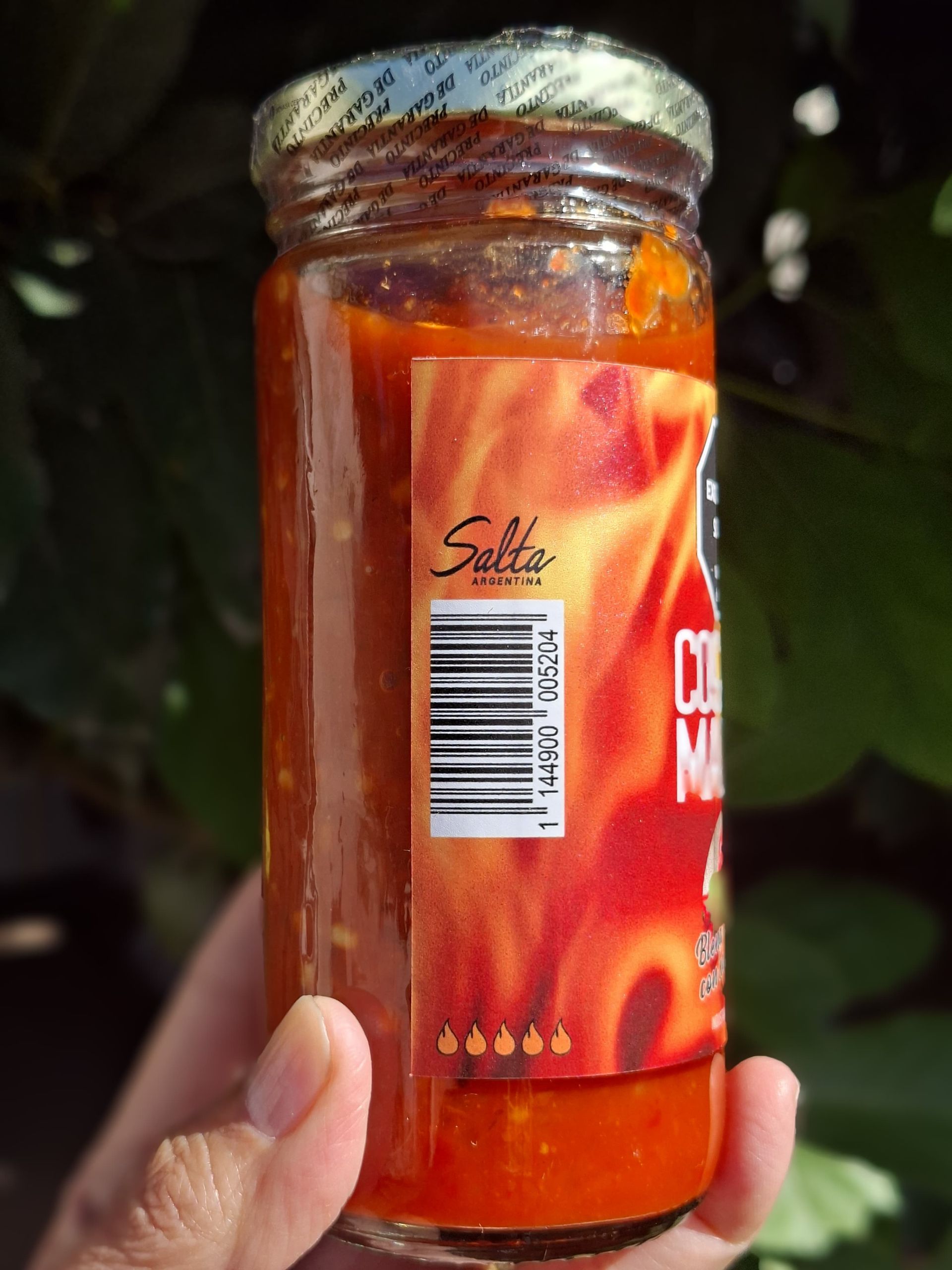 Mano sosteniendo un frasco de salsa especiada de color rojo anaranjado con etiqueta y código de barras.