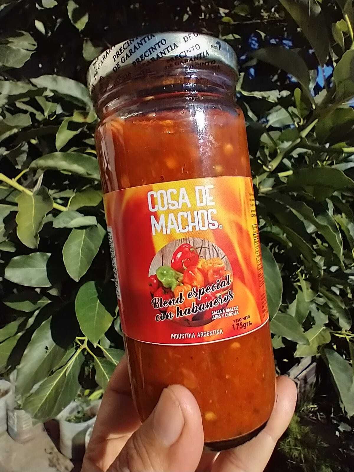 Una mano sostiene un frasco de salsa de naranja con una etiqueta roja, al aire libre, con hojas verdes de fondo.