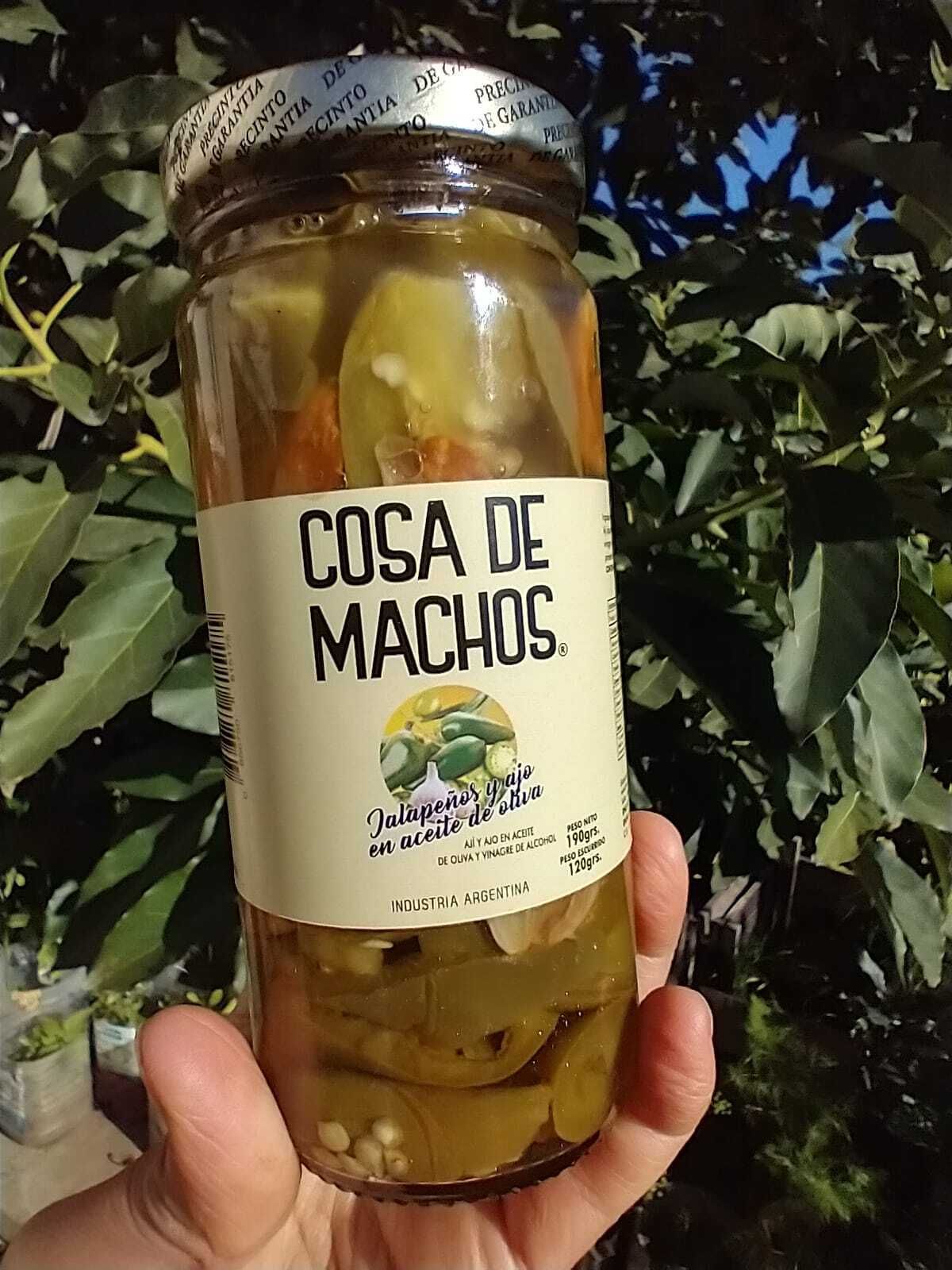 Frasco etiquetado como “COSA DE MACHOS”, colocado al aire libre, con pimientos encurtidos en líquido color ámbar.