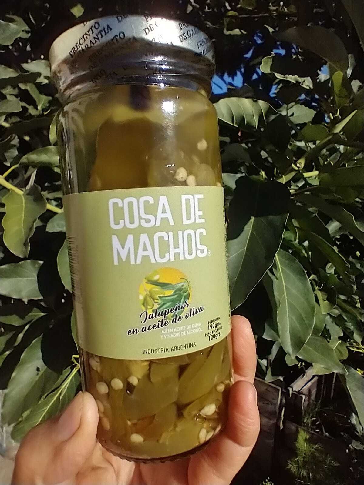Una mano sostiene un frasco con la etiqueta “COSA DE MACHOS” frente a plantas de hojas verdes.