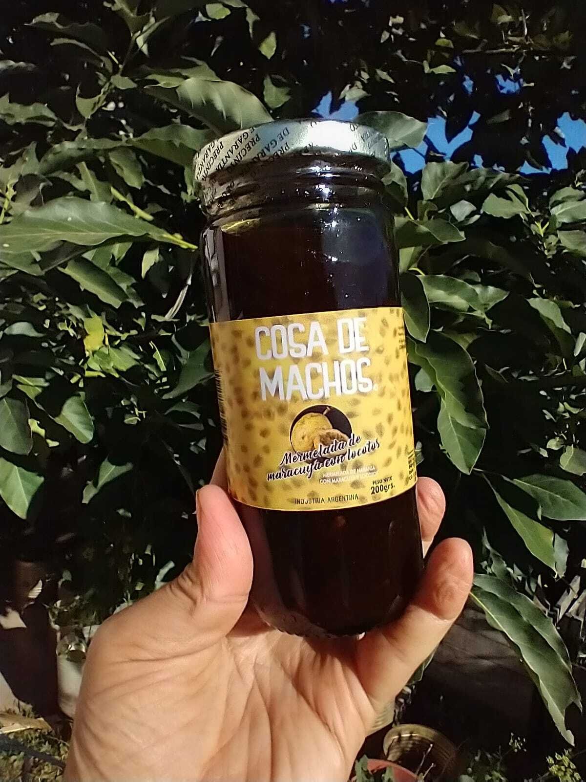 Una mano sostiene un frasco etiquetado como “Coca de Macha” al aire libre, frente a plantas de hojas verdes.