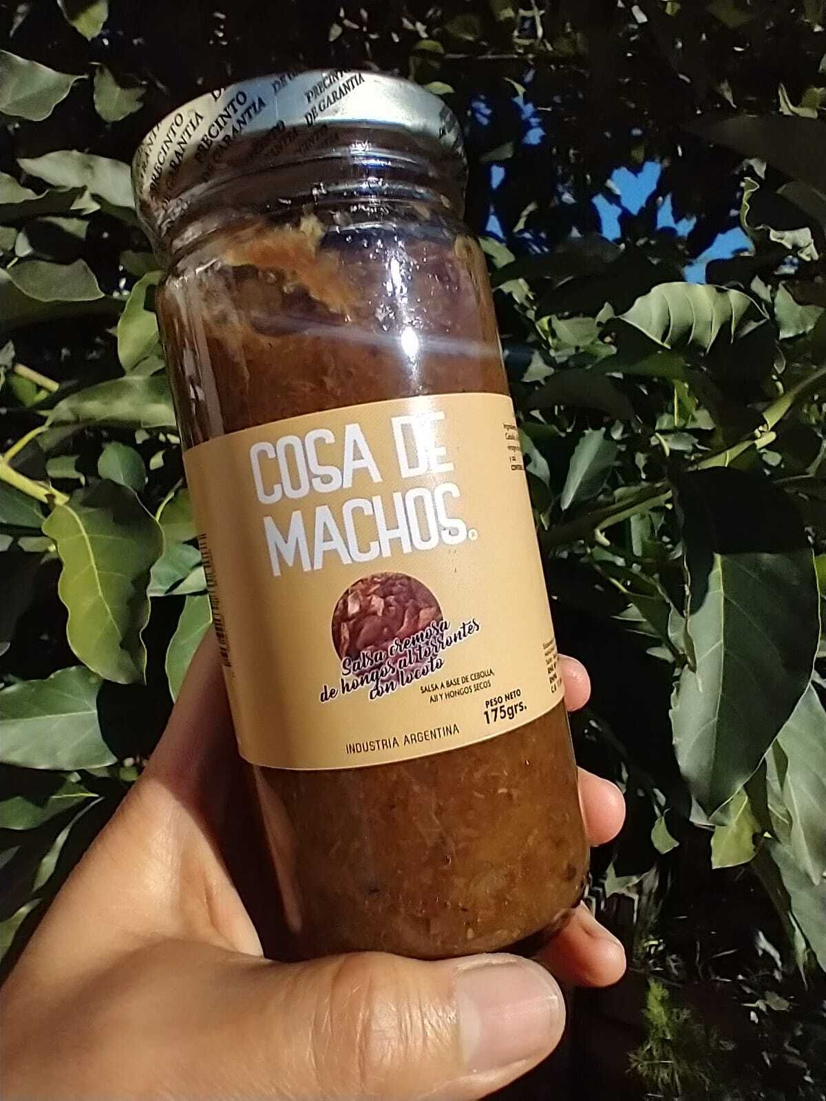 Una mano sostiene un frasco etiquetado como “Cosa de Machos” al aire libre, con el follaje verde de fondo.