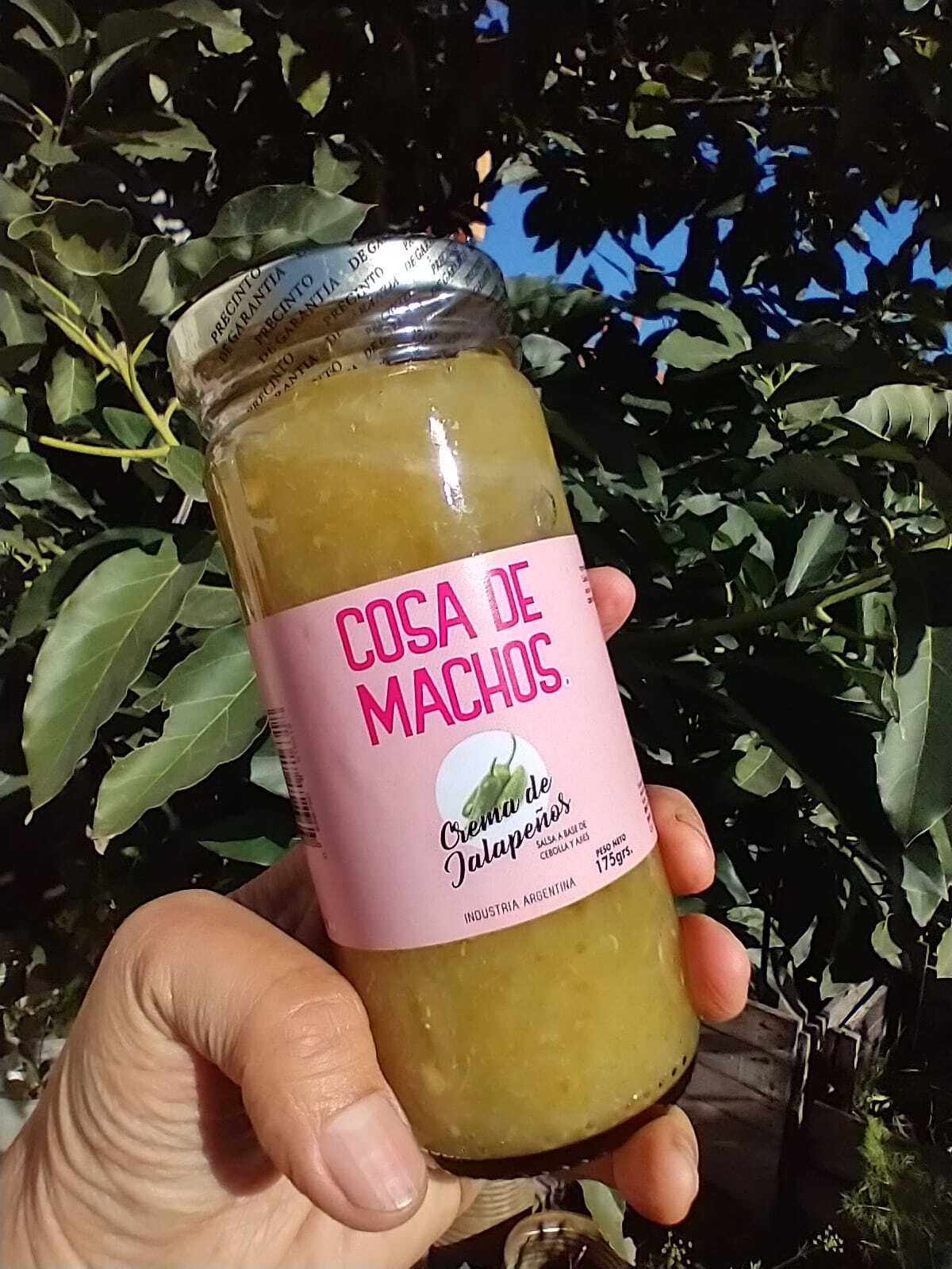 Una mano sostiene un pequeño frasco con la etiqueta “Coca de Machos” al aire libre, con vegetación frondosa de fondo.