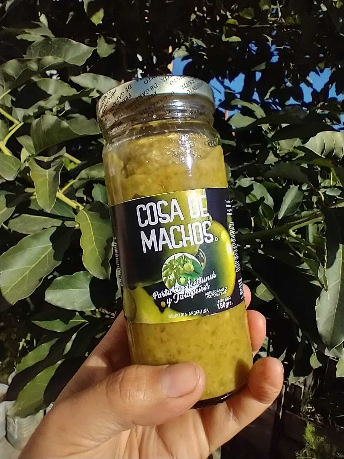 Una mano sostiene un frasco de salsa amarilla con la etiqueta “Cosa Machos” frente a plantas de hojas verdes.
