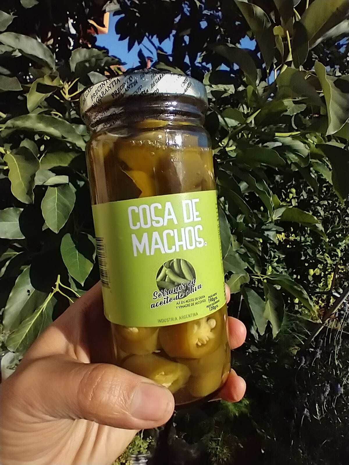 Una mano sostiene un frasco de pimientos encurtidos con etiqueta verde al aire libre, apoyado contra ramas frondosas.